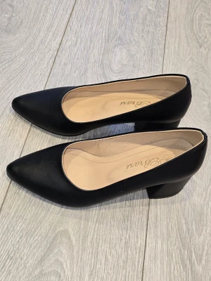 Svarta pumps med låg klack från Bravi - Stilrena svarta pumps från Bravi med spetsig tå och låg, bred klack. Skorna är tillverkade i slätt konstläder med beige innersula för extra komfort. Perfekta för dig som vill ha en klassisk och tidlös look.