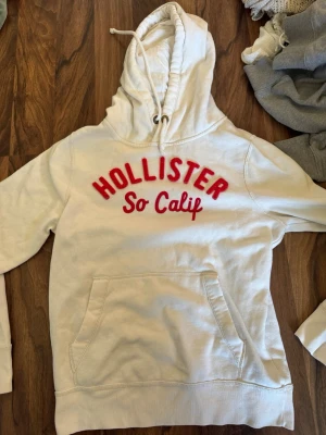 Vit hoodie från Hollister med röd text - Säljer en vit hoodie från Hollister med röd broderad text 'HOLLISTER So Calif' på bröstet. Hoodien har en stor känguruficka framtill, dragsko i huvan och mjukt fleecefoder på insidan. Perfekt för en avslappnad och chill stil.