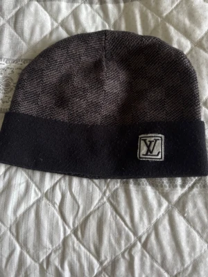 Mörkgrå L V mössa - Snygg mörkgrå mössa, med diskret rutmönster och svart uppvikt kant. På kanten sitter en broderad logga i vitt och svart. Perfekt accessoar för att lyfta din stil under kyliga dagar.
