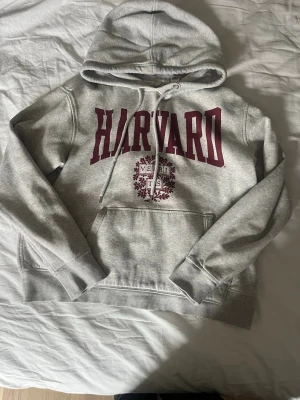 Grå Harvard hoodie från H&M - Snygg grå hoodie från H&M med stor vinröd HARVARD-text och emblem på bröstet. Klassisk känguruficka framtill, ribbade muddar och justerbar huva med dragsko. Perfekt för en avslappnad och cool stil.