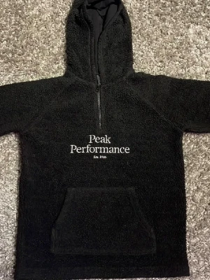 Svart teddytröja Peak Performance - Mysig svart teddytröja från Peak Performance med huva och halv dragkedja framtill. Stor magficka och broderad logga på bröstet. Perfekt för kalla dagar och riktigt skön att ha på sig. Materialet är fluffigt och värmande.