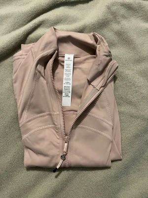 Beige zip-jacka från Lululemon - Snygg och figursydd beige zip-jacka från Lululemon med hög krage och långa ärmar. Jackan har praktiska fickor med dragkedja och är tillverkad i ett stretchigt och mjukt material som passar perfekt för träning eller sport. Diskreta sömmar ger en stilren look.