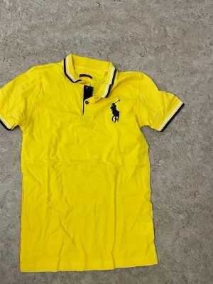 Gul pikétröja från Ralph Lauren - Gul pikétröja från Ralph Lauren med klassisk krage och knappslå. Tröjan har korta ärmar och svart broderad logga på bröstet. Snygga svarta detaljer på krage och ärmslut. Perfekt för en sportig och stilren look.