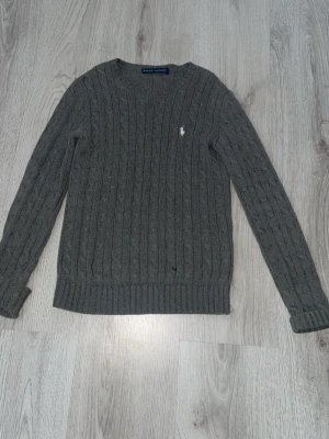 Grå kabelstickad tröja Ralph Lauren - Kabelstickad grå tröja från Ralph Lauren med rund halsringning och klassisk vit broderad logga på bröstet. Tröjan har ribbade muddar vid ärmslut och nederkant, perfekt för en stilren och tidlös look.