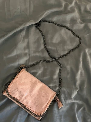Rosa axelväska med kedjerem - Snygg axelväska i ljusrosa mockaliknande material med dekorativa svarta kedjedetaljer runt kanterna och som axelrem. Väskan har magnetlås och flera fack inuti för småsaker. Perfekt för dig som gillar trendiga accessoarer med lite edge.