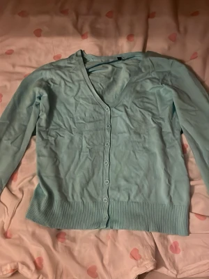 Ljusblå cardigan med v-ringning - Snygg ljusblå cardigan med v-ringning och knappar hela vägen framtill. Tröjan har långa ärmar och bred ribbstickad mudd vid ärmslut och nederkant. Perfekt att slänga över en t-shirt eller linne för en chill look.