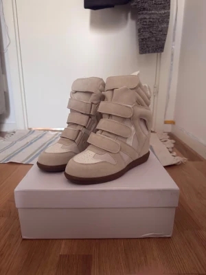 Beige Isabel Marant - Snygga beige sneakers med inbyggd kilklack och flera breda kardborreband. Skorna har en mix av mocka och textil, samt en brun sula. Perfekta för dig som vill ha både höjd och streetstyle-vibe i ett par skor.