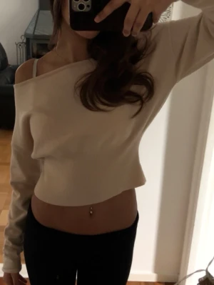Beige offshoulder croppad topp - Trendig beige croppad topp med offshoulder-design och långa ärmar. Toppens material är ribbad och ger en snygg passform som framhäver midjan. Perfekt för dig som gillar en avslappnad men ändå stilren look.