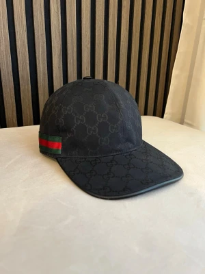 Svart keps GG - Snygg svart keps från Gucci med klassiskt GG-monogram över hela kepsen. På sidan finns en ikonisk grön och röd rand. Justerbar rem baktill för perfekt passform. Perfekt accessoar för en trendig streetstyle-look.