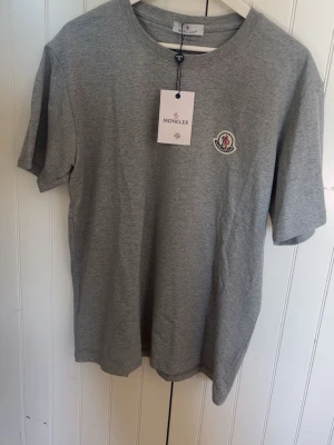 Grå Moncler t-shirt med logga - Snygg grå t-shirt från Moncler med klassisk rund hals och broderad logga på bröstet. T-shirten är kortärmad och tillverkad i mjuk bomull, perfekt för en clean och stilren look.
