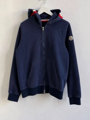 Marinblå hoodie från Moncler - Snygg marinblå hoodie från Moncler med dragkedja framtill och klassisk huva. Fodret i huvan har rödvita detaljer och ärmen pryds av Monclers logotyp. Hoodien har känguruficka och ribbade muddar vid ärmslut och nederkant. Perfekt för en avslappnad stil.