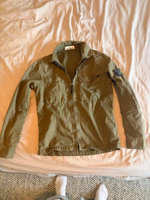Olivgrön overshirt från Stone Island - Snygg olivgrön overshirt från Stone Island med dragkedja framtill, två bröstfickor och klassisk patch på vänster ärm. Priset är ej hugget i sten