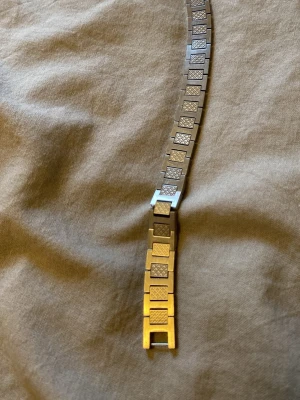 Tvåfärgad länkarmband till klocka - Snyggt länkarmband till klocka i både silver och guldton. Armbandet har rektangulära länkar med matta och blanka ytor, samt ett diskret rutmönster på vissa länkar. Passar dig som vill ha en stilren och modern look på din klocka.