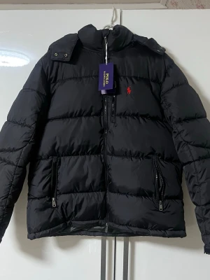 Svart pufferjacka från Polo Ralph Lauren - Svart pufferjacka från Polo Ralph Lauren med röd logga på bröstet, två stora fickor med dragkedja och hög krage. Jackan har en klassisk pufferdesign och blanka dragkedjor med Polo-gravyr. Perfekt för kalla vinterdagar.