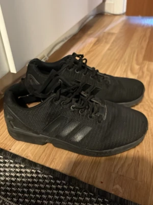 Svarta Adidas sneakers i mesh - Säljer ett par helsvarta Adidas sneakers med lågt skaft. Skorna har en stilren design med tre svarta ränder på sidan och ovandel i meshmaterial för bra ventilation. Yttersulan är robust och snörningen klassisk. Perfekta för dig som gillar sportig och clean stil.