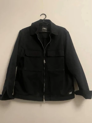 Svart overshirt från ZARA - Stilren svart overshirt från ZARA med dragkedja framtill och två stora bröstfickor med lock. Overshirten har klassisk krage och raka ärmar, perfekt för lager-på-lager. Materialet är kraftigt och ger en snygg siluett.