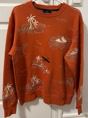 Orange tröja  - Cool orange hoodie med broderade motiv av palmer, vågor och surfbräda. Rund halsringning och ribbade muddar. Perfekt för dig som gillar unika detaljer och avslappnad stil.
