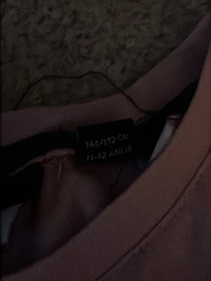 Rosa GANT t-shirt med tryck - Säljer en rosa t-shirt från GANT med svart logotyp och sköldtryck på bröstet. Klassisk rund halsringning och korta ärmar. Perfekt för dig som gillar stilrena och sportiga plagg. Materialet känns mjukt och skönt mot huden.