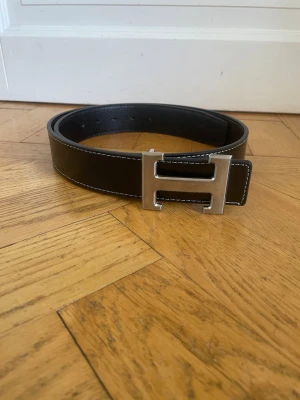 H-bälte - Stilrent svart skärp med ikoniskt H-format spänne i silverfärgad metall. Skärpet har vita kontrastsömmar längs kanterna och är tillverkat i äkta skinn. Perfekt accessoar för att lyfta dina outfits. 