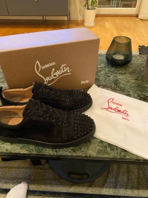 Svarta Christian Louboutin sneakers - Snygga svarta sneakers från Christian Louboutin i mocka med coola svarta nitar på tån. Klassisk låg modell med svarta skosnören och diskret logga på insidan. Perfekta för dig som vill sticka ut med en lyxig och edgy stil.