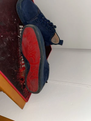 Blåa sneakers från Christian Louboutin - Mörkblå sneakers i mocka från Christian Louboutin med klassisk röd sula och broderad logga på hälen. Skorna har lågt skaft, blåa skosnören och en stilren design som sticker ut med den ikoniska röda undersidan.
