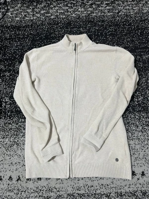 Beige stickad ziptröja - Snygg beige stickad ziptröja med låg krage och hel dragkedja framtill. Tröjan har ribbade muddar vid ärmslut och nederkant samt en liten metallogo nertill. Perfekt för lager på lager och enkel att matcha. De där små fläckarna på ryggen går bort med tvätt 