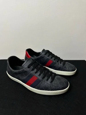 Gucci Ace  - Snygga Gucci Ace sneakers i svart läder med klassiska röda och mörkblå ränder på sidorna. Skorna har diskret GG-mönster över hela ovandelen och vit sula. Röd detalj vid hälen ger extra stilpoäng. Perfekta för dig som vill sticka ut med lyxig streetstyle.