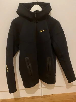 Svart Nike NOCTA hoodie med dragkedja - Svart hoodie från Nike NOCTA med hel dragkedja, två fickor med dragkedja och gul logga på bröstet. NOCTA-tryck på ärmen. Huvan är stor och ger en cool streetwear-look. Perfekt för dig som gillar stilrena och sportiga plagg.
