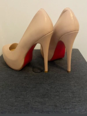 Beige pumps med röd sula - Snygga pumps i beige läder med klassisk spetsig tå och hög stilettklack. Skorna har den ikoniska röda sulan som ger en lyxig känsla. Perfekta för dig som vill sticka ut med en elegant och tidlös look.