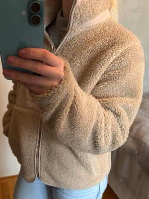 Beige teddyjacka med dragkedja - Mysig beige teddyjacka med hög krage och hel dragkedja framtill. Jackan har en bröstficka med dragkedja i matchande beige tyg och är tillverkad i fluffigt teddy-material. 