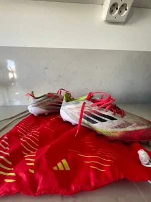 Adidas X Speedflow fotbollsskor - Säljer ett par Adidas X Speedflow fotbollsskor i vitt, rött och neongult med svarta detaljer. Skorna har snörning och en lätt, ventilerande ovandel i syntetmaterial. Medföljer en röd Adidas-påse med gula detaljer. Perfekta för snabba spelare på planen.