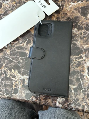 Svart mobilskal från Holdit med magnet iPhone 11 Pro Max/xs Max  - Snyggt svart mobilskal i plånboksmodell från Holdit. Fodralet är gjort i veganskt läder och har magnetstängning. Man kan ta av fodralet som ni ser på 3de bilden och ha andra mobilskalen för sig själv, då den är magnetisk. För iPhone 11 Pro Max /Xs Max 