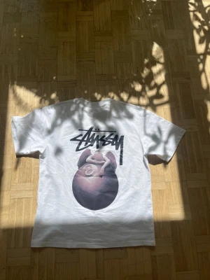 Vit Stüssy t-shirt  - Vit t-shirt från Stüssy med stort grafiskt tryck på ryggen och mindre tryck på bröstet. Klassisk passform med rund hals och korta ärmar. Perfekt för dig som gillar streetwear och vill sticka ut med en unik design.