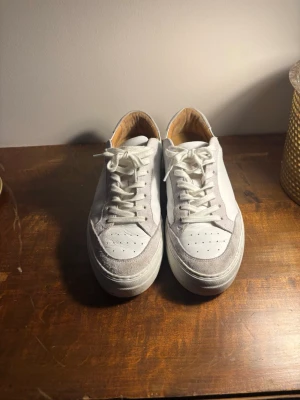 Vita sneakers med mockadetaljer/ j.linderberg  - Snygga vita sneakers med grå mockadetaljer runt tån och hälen. Skorna har klassisk låg profil, vita skosnören och perforerad tå för extra ventilation. Ovandelen är i läder och mocka, vilket ger en stilren och fräsch look.