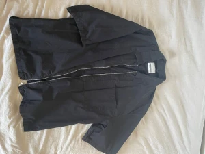 Svart overshirt med dragkedja - Stilren svart overshirt med korta ärmar och dragkedja framtill. Overshirten har en stor bröstficka och klassisk krage. Enkel och clean design som passar till det mesta.