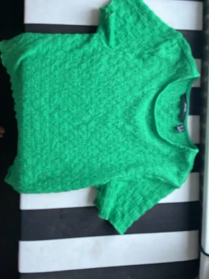 Grön kortärmad topp från H&M - Snygg grön topp från H&M med korta ärmar och en texturerad yta. Modellen är croppad och har en rund halsringning. Perfekt att styla med jeans eller kjol för en trendig look.