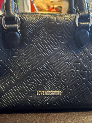 Svart handväska från Love Moschino - Snygg svart handväska från Love Moschino med präglad logotyp och hjärtdetaljer över hela väskan. Väskan har dubbla handtag med silvriga metalldetaljer och är tillverkad i konstläder. Perfekt för dig som gillar trendiga accessoarer. Dragkedja behövs lagas