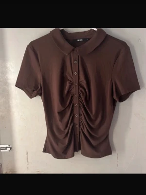 Brun topp  - Snygg brun topp från Bik Bok med korta ärmar, klassisk krage och knappar hela vägen framtill. Toppen har en rynkad detalj längs knappraden som ger en cool och trendig look. Perfekt att styla med jeans eller kjol. Perfekt till sommar!