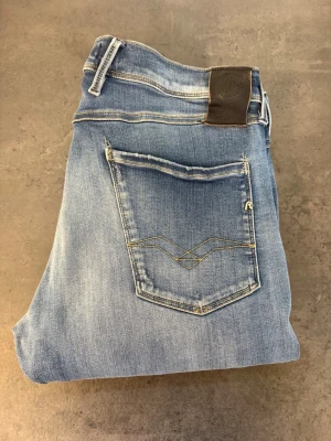Blå slim fit jeans Replay Anbass W34/L33 - Snygga blå slim fit jeans från Replay med klassisk femficksdesign och ljusa slitningar på låren. Jeansen har en läderpatch med logga bak i midjan och dekorativa sömmar på bakfickan. Perfekta för en avslappnad och trendig look.