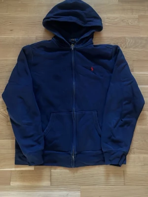 Mörkblå hoodie från Polo Ralph Lauren - Mörkblå hoodie med dragkedja från Polo Ralph Lauren. Klassisk design med huva, kängurufickor och röd broderad logga på bröstet. Perfekt för en avslappnad och stilren look.