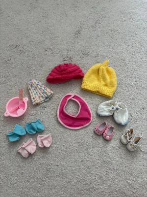Dockkläder och accessoarer mix - Säljer ett set med dockkläder och accessoarer: tre stickade mössor i gult, rött och flerfärgat, 1 haklapp i rosa, fyra par sockor i blått och rosa, tre par skor i vitt och rosa samt en rosa matskål med sked. Perfekt för att styla dockan!