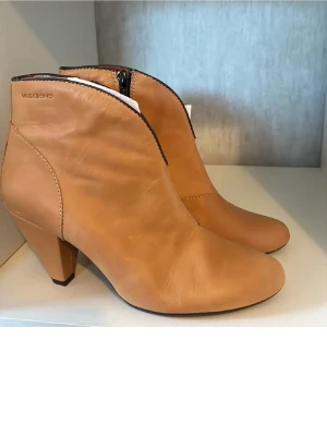Beiga skinnboots från Vagabond - Snygga boots från Vagabond i beige skinn med rund tå och låg klack. Skorna har dragkedja på sidan och en stilren design med diskreta sömmar. Insidan är fodrad i rött för en cool kontrast. Perfekta för dig som gillar klassisk men trendig stil. Obs liten prick/fläck på ena sidan se bild. Inget jag tänker på. Säg till för fler bilder