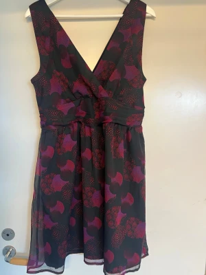 Svart och lila klänning XL Vero Moda  - Snygg klänning med svart bas och lila samt röda blommiga mönster. Klänningen har v-ringning, markerad midja och är ärmlös. Perfekt för dig som vill ha en feminin och stilren look.