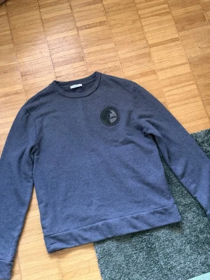 Mörkgrå Versace sweatshirt  - Säljer en mörkgrå sweatshirt med rund hals och ett svart tryck med logga på bröstet. Tröjan har långa ärmar och ribbade muddar vid ärmslut och nederkant. Perfekt för en chill och avslappnad stil.
