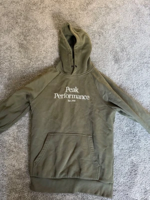 Olivgrön hoodie Peak Performance - Snygg olivgrön hoodie från Peak Performance med stor magficka och broderad logga på bröstet. Klassisk huva med metallöljetter och mjukt material på insidan. Perfekt för en avslappnad och sportig stil.