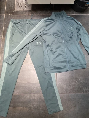 Under armour tracksuit - Snyggt ljusblått träningsset från Under Armour med matchande zip-jacka och byxor. Setet har ljusare blå sidopaneler och logga på både jacka och byxa. Tillverkat i mjukt funktionsmaterial, perfekt för träning eller chill.