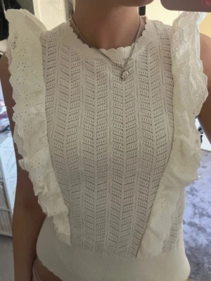 Vit ärmlös topp med spetsvolanger - Supersöt vit ärmlös topp med broderade spetsvolanger längs axlarna. Stickad struktur med sicksackmönster och rund halsringning. Perfekt för en trendig och feminin look.