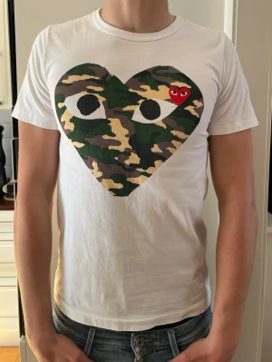 Vit Comme des Garçons t-shirt med camo-hjärta - Vit t-shirt från Comme des Garçons Play med stort hjärta i camouflage-mönster och ett litet rött hjärta på bröstet. Klassisk passform och rund halsringning. Perfekt statement-plagg för dig som gillar streetwear. Är M men passar S. Lite gul fläckig i ena armhålan där av priset.
