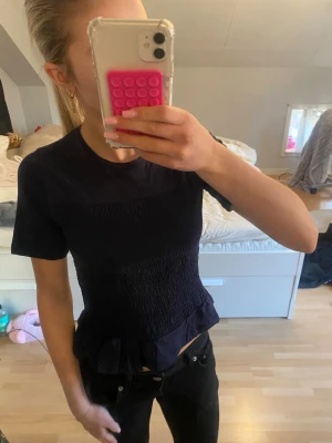 Mörkblå topp från Gina Tricot - Snygg mörkblå topp från Gina Tricot med korta ärmar och smockad midja som ger en figurnära passform. Toppen har en liten volangkant nedtill och är tillverkad i mjuk bomull. Perfekt att styla med jeans eller kjol.