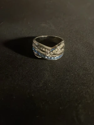 Silverfärgad ring med blå stenar - Snygg silverfärgad ring med flera band som korsar varandra, dekorerad med små klara och blå stenar för extra bling. Ringen har en modern och unik design som verkligen sticker ut.OBS! Materialet är okänt, kan ej garantera att det är rostfritt.